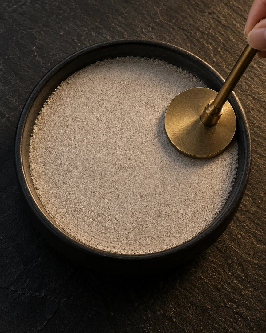 A brass ash press smoothing fine pale ash in a black ceramic dish
